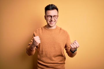 Young handsome caucasian man wearing glasses and casual winter sweater over yellow background excited for success with arms raised and eyes closed celebrating victory smiling. Winner concept.