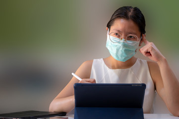 Asian woman wearing surgical mask thinking and working by using tablet isolated on blurred...