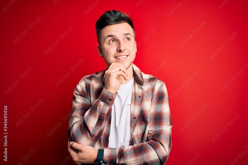 Wall mural Young handsome caucasian man wearing casual modern shirt over red isolated background with hand on chin thinking about question, pensive expression. Smiling and thoughtful face. Doubt concept.