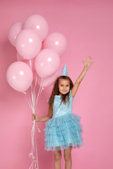 adorable smiling little child girl in blue dress and birthday hat celebrating with pastel pink air balloons on pink background. birthday party.