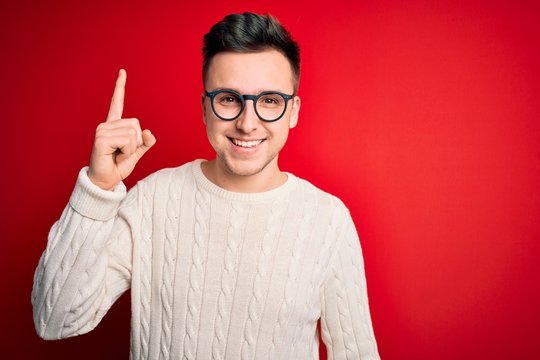Young handsome caucasian man wearing glasses and casual winter sweater over red background pointing finger up with successful idea. Exited and happy. Number one.