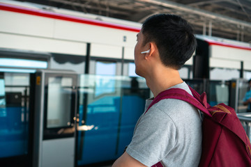 Close up of young man listening to music with wireless earpods while commuting by train at station. Asian guy enjoying music on the go.