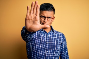 Young handsome latin man wearing casual shirt and glasses over yellow background doing stop sing...