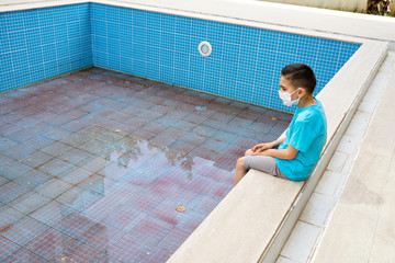 A boy with medical mask sits by an empty pool. Concept summer 2020. No tourism 