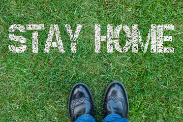 Black shoes standing next to text stay home. Text stay home painted on green grass.