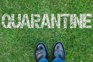 Black shoes standing next to text quarantine. Text quarantine painted on green grass.