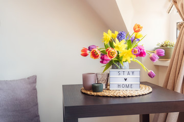 Everything for a calm stay at home. Lightbox with Stay home message, bouquet of fresh spring flowers and candles on the coffee table, and pillow sitting place. Cozy area for relaxing near window.