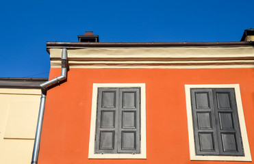 Fototapeta na wymiar Red painted wall with two windows and closed shutters under blue sky in historic part of Lviv city, Ukraine