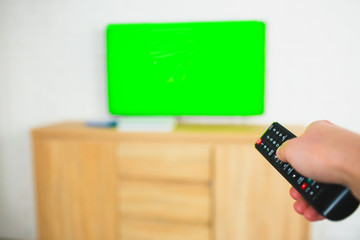 Young man sit in room during quarantine. Guy hold remote control in hand and applying it to tv with green screen. Modern technologies and super device.
