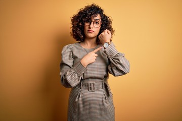 Beautiful arab business woman wearing dress and glasses standing over yellow background In hurry...