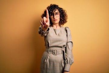 Beautiful arab business woman wearing dress and glasses standing over yellow background Pointing with finger up and angry expression, showing no gesture