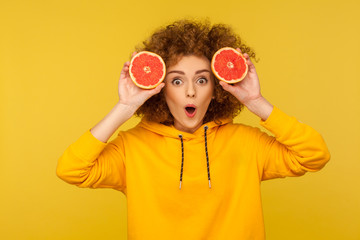 Surprised lively energetic woman with fluffy curly hairstyle holding half slice grapefruit near face, looking at camera with amazement, healthy raw fresh food concept and beauty care. studio shot
