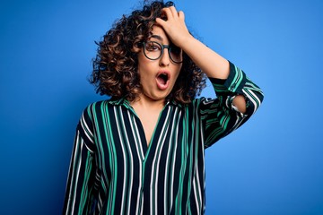 Young beautiful curly arab woman wearing striped shirt and glasses over blue background surprised...