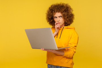 Confused puzzled thoughtful woman with fluffy curly hair in urban style hoody holding laptop...