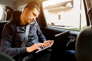 Busy businessman in a taxi. Multitasking concept. Passenger rides in the back seat and works simultaneously. Speaks on a smartphone and uses a laptop and tablet.