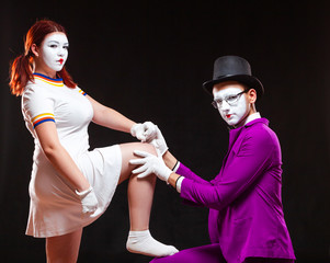 Portrait of two mime artists, isolated on black background. Young woman puts her foot on mans knee....