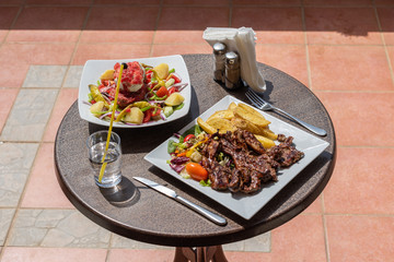 Lunch on the plate with lamb, potato and salad on the table outside on the balcony. Spring or summer blooming trees and flowers in the garden.