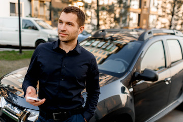 A young man or businessman in a dark shirt stands on the street near the car, looks into the distance in a residential area of the city. The driver is waiting for his passenger or client.