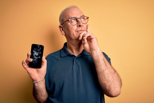 Middle age man holding broken smartphone showing cracked screen over yellow background serious face thinking about question, very confused idea