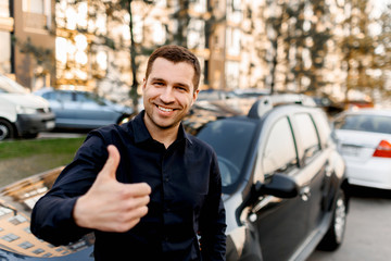 A young man stands near a car and shows a thumb up.Taxi driver looks at the camera and smiles....