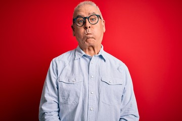 Middle age handsome hoary man wearing casual striped shirt and glasses over red background puffing cheeks with funny face. Mouth inflated with air, crazy expression.