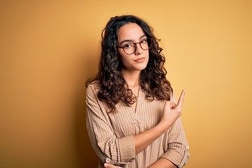 Beautiful woman with curly hair wearing striped shirt and glasses over yellow background Pointing with hand finger to the side showing advertisement, serious and calm face