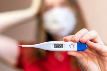 Girl in a medical mask demonstrates an Digital thermometer. Concept of illness, fever. Selective focus, blurred background.