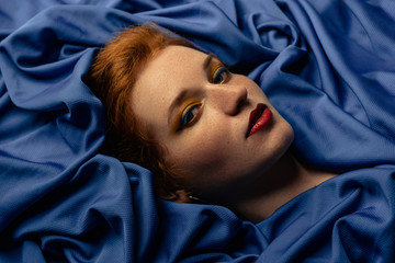 Beautiful redhead freckled woman with colorful makeup laying on dark blue sheets. Close up studio...