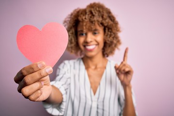Young african american romantic woman with curly hair holding paper heart shape surprised with an idea or question pointing finger with happy face, number one