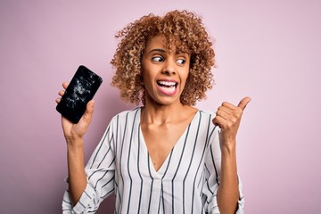 Young african american curly woman holding broken smartphone showing craked screen pointing and...