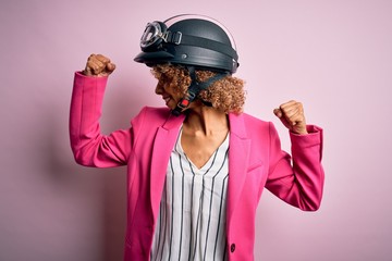 African american motorcyclist woman with curly hair wearing moto helmet over pink background showing arms muscles smiling proud. Fitness concept.
