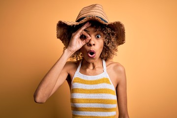 African american tourist woman with curly on vacation wearing summer hat and striped t-shirt doing ok gesture shocked with surprised face, eye looking through fingers. Unbelieving expression.