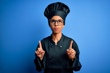 Young african american chef woman wearing cooker uniform and hat over blue background Pointing up looking sad and upset, indicating direction with fingers, unhappy and depressed.