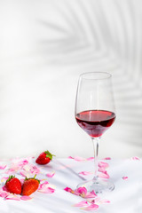A glass of red wine with petals and some strawberries on a white background, with palm shadows and copy space for text