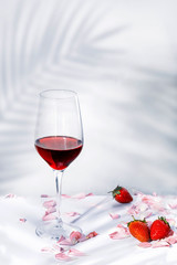 A glass of red wine with petals and some strawberries on a white background, with palm shadows and copy space for text