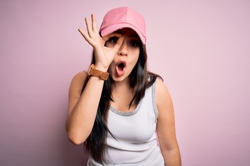 Young brunette woman wearing casual sport cap over pink background doing ok gesture shocked with surprised face, eye looking through fingers. Unbelieving expression.