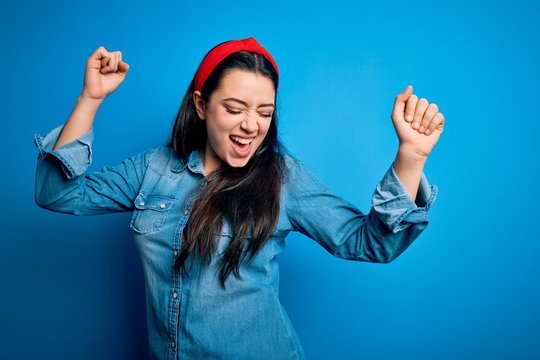 Young brunette woman wearing casual denim shirt over blue isolated background Dancing happy and cheerful, smiling moving casual and confident listening to music