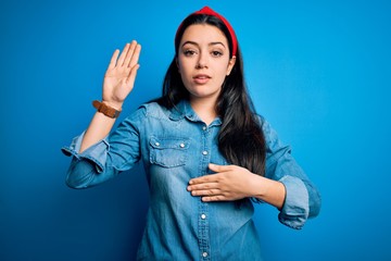 Young brunette woman wearing casual denim shirt over blue isolated background Swearing with hand on...
