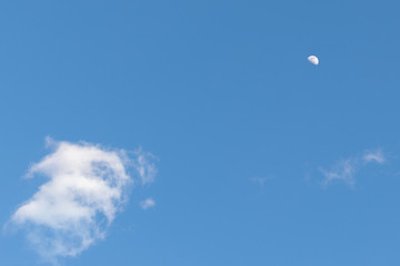 The half moon surround by clouds in the bright blue daytime sky.