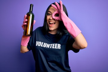 Young brunette woman wearing volunteer t-shirt picking glass bottle over purple background with happy face smiling doing ok sign with hand on eye looking through fingers