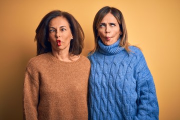 Middle age beautiful couple of sisters wearing casual sweater over isolated yellow background making fish face with lips, crazy and comical gesture. Funny expression.