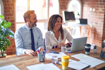 Middle age beautiful business workers working together using laptop at the office looking away to side with smile on face, natural expression. Laughing confident.