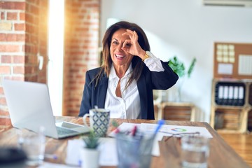 Middle age beautiful businesswoman working using laptop at the office doing ok gesture with hand smiling, eye looking through fingers with happy face.