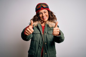 Middle age skier woman wearing snow sportswear and ski goggles over white background approving doing positive gesture with hand, thumbs up smiling and happy for success. Winner gesture.