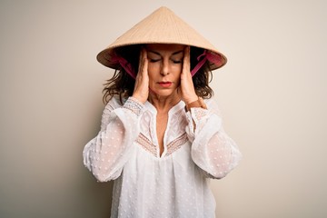 Middle age brunette woman wearing asian traditional conical hat over white background suffering from headache desperate and stressed because pain and migraine. Hands on head.