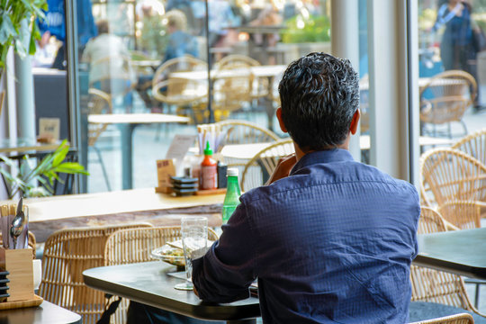 Man from behind sits alone in a modern cafe restaurant and looks outside. He is waiting for his companion to return. There is no one else around him. Social distancing.