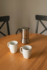 coffee morning at home vertical close-up; two white coffee mugs and a coffee maker over a wooden table with black chairs and a white wall