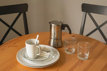 after breakfast morning at home close-up; two white coffee mugs, a metal coffee maker, empty white dishes, and glasses over a wooden table with black chairs
