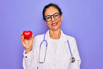 Middle age senior cardiologist doctor woman holding red heart over purple background with a happy...