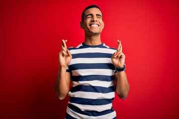 Handsome african american man wearing casual striped t-shirt standing over red background gesturing finger crossed smiling with hope and eyes closed. Luck and superstitious concept.